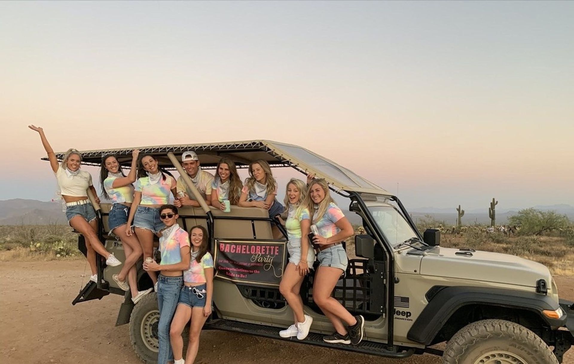 Group of friends in tie-dye shirts posing on an open-air tour Jeep at sunset in a desert with saguaro cacti for a bachelorette party.