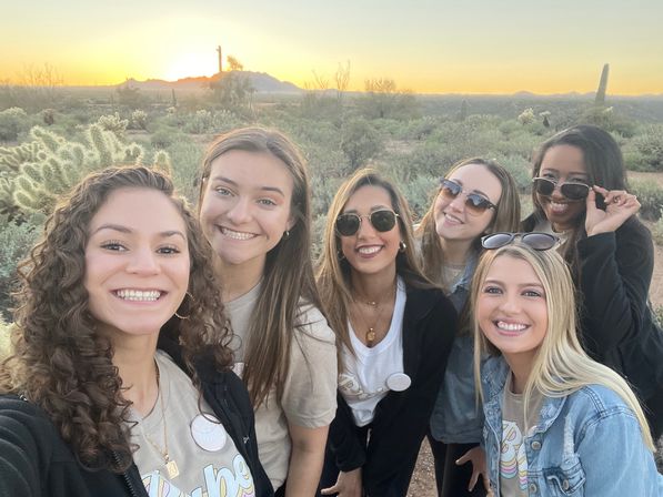 Six friends taking a smiling group selfie at sunset in a cactus-filled desert with saguaro silhouettes and distant mountains