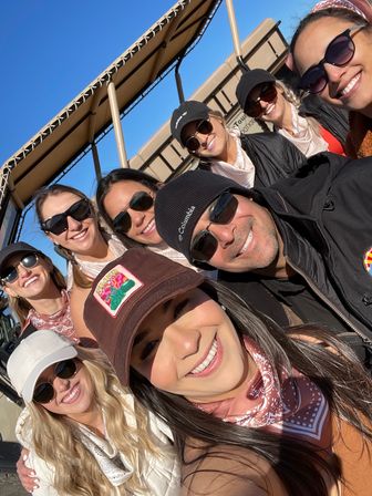 Smiling group selfie of eight people in sunglasses, caps and bandanas riding an open-air tour vehicle under a clear blue sky on a sunny outdoor excursion.