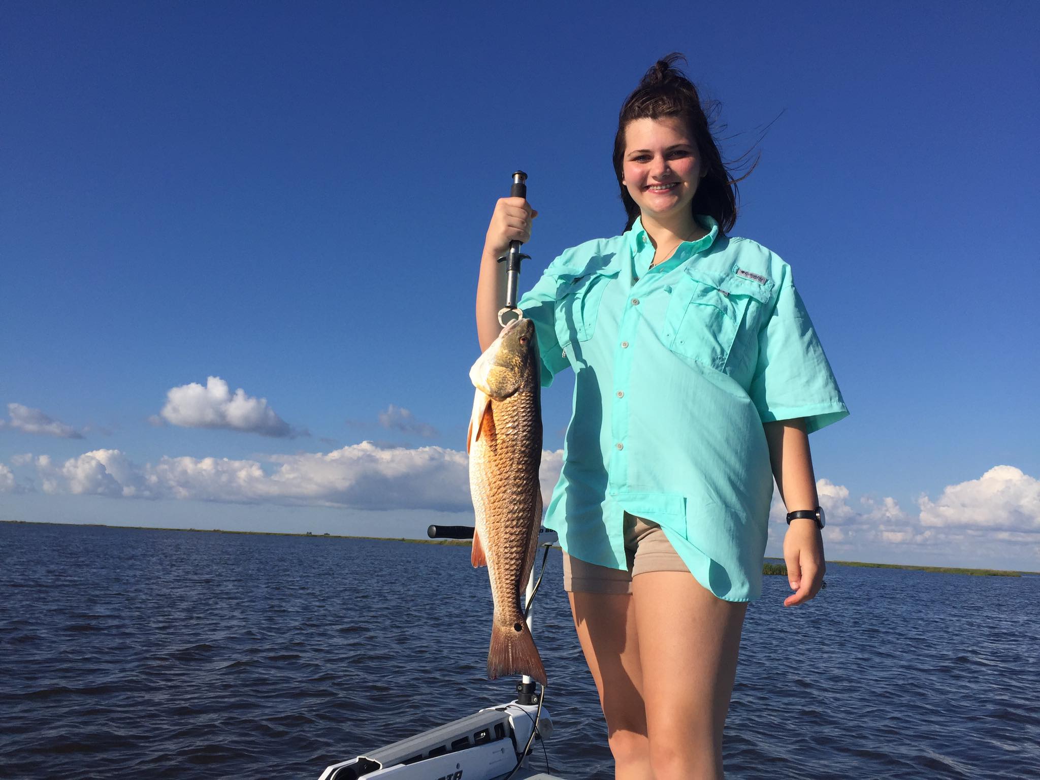 Smiling inshore angler on a small boat holding a large redfish with a fish gripper over calm coastal bay waters under a bright blue sky with scattered clouds.