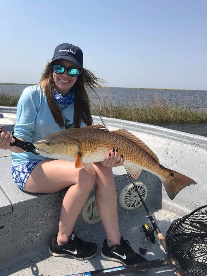 Smiling angler on a skiff holding a large redfish (red drum) in a shallow bay near marsh grasses, with fishing rod and net visible on the boat deck.