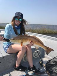 Smiling angler on a skiff holding a large redfish (red drum) in a shallow bay near marsh grasses, with fishing rod and net visible on the boat deck.