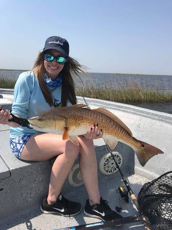 Smiling angler on a skiff holding a large redfish (red drum) in a shallow bay near marsh grasses, with fishing rod and net visible on the boat deck.