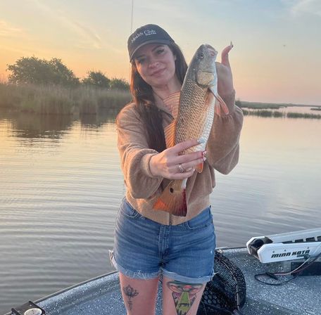 Person on a boat at sunset holding a freshly caught redfish over calm coastal marsh waters — casual sweater, denim shorts and visible leg tattoos, catch-and-release fishing scene.