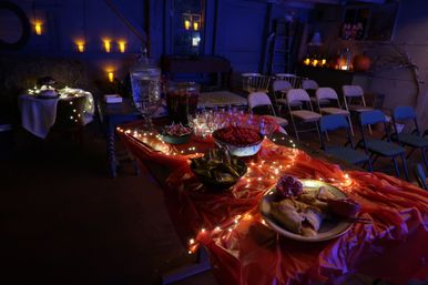 Dimly lit rustic barn party with a red-draped buffet table wrapped in fairy lights, pitchers and bowls of snacks, rows of folding chairs and candlelit pumpkins in the background.