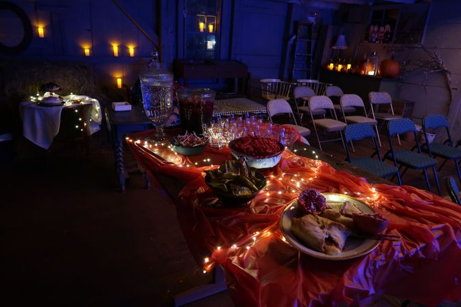 Dimly lit rustic barn party with a red-draped buffet table wrapped in fairy lights, pitchers and bowls of snacks, rows of folding chairs and candlelit pumpkins in the background.