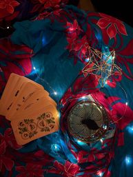 Boho mystic reading setup with fanned yellow oracle/tarot cards, a teacup showing tea leaves for tasseography, a gold wire star ornament and warm fairy lights on bright blue-and-pink floral fabric.