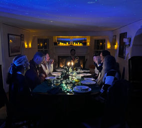 People laughing around a candlelit dinner table set with plates, glassware and string-light centerpiece, a fireplace lined with flickering candles and a blue starry ceiling projection creating a cozy, magical atmosphere.