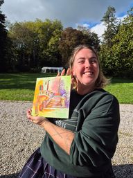Smiling person outdoors on a sunny day holding a small vibrant painting of a stairway; green lawn, tall trees and a white canopy in the background with gravel in the foreground.