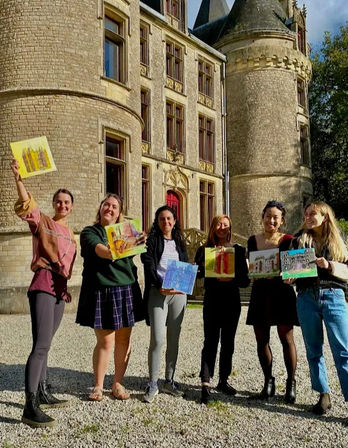 Six people smiling in a gravel courtyard in front of a historic stone chateau, each proudly holding a small colorful painting from a sunny plein-air art session.