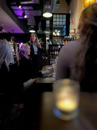 Group of women enjoying wine at a cozy urban wine bar, candlelit wooden table, shelves of bottles and pendant lights overhead
