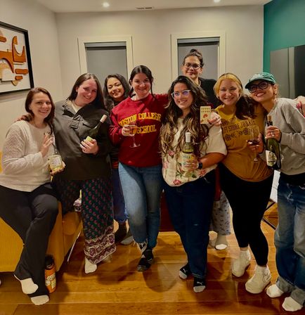 Eight friends smiling in a cozy living room for an at-home wine night, holding bottles and glasses on hardwood floors
