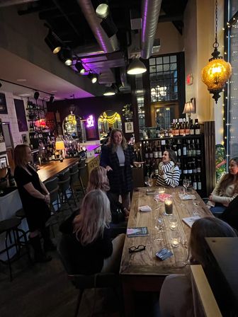 Cozy city wine bar interior at night with a long wooden communal table, candles and empty wine glasses, a small group chatting, neon signs and warm pendant lighting