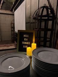 Rustic indoor event setup with a letter board reading 'HELL IS A TEENAGE GIRL', glowing LED candles, stacked white plates, vintage metal cage lanterns and a projection screen in a dimly lit barn-style venue.