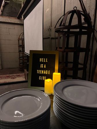 Rustic indoor event setup with a letter board reading 'HELL IS A TEENAGE GIRL', glowing LED candles, stacked white plates, vintage metal cage lanterns and a projection screen in a dimly lit barn-style venue.