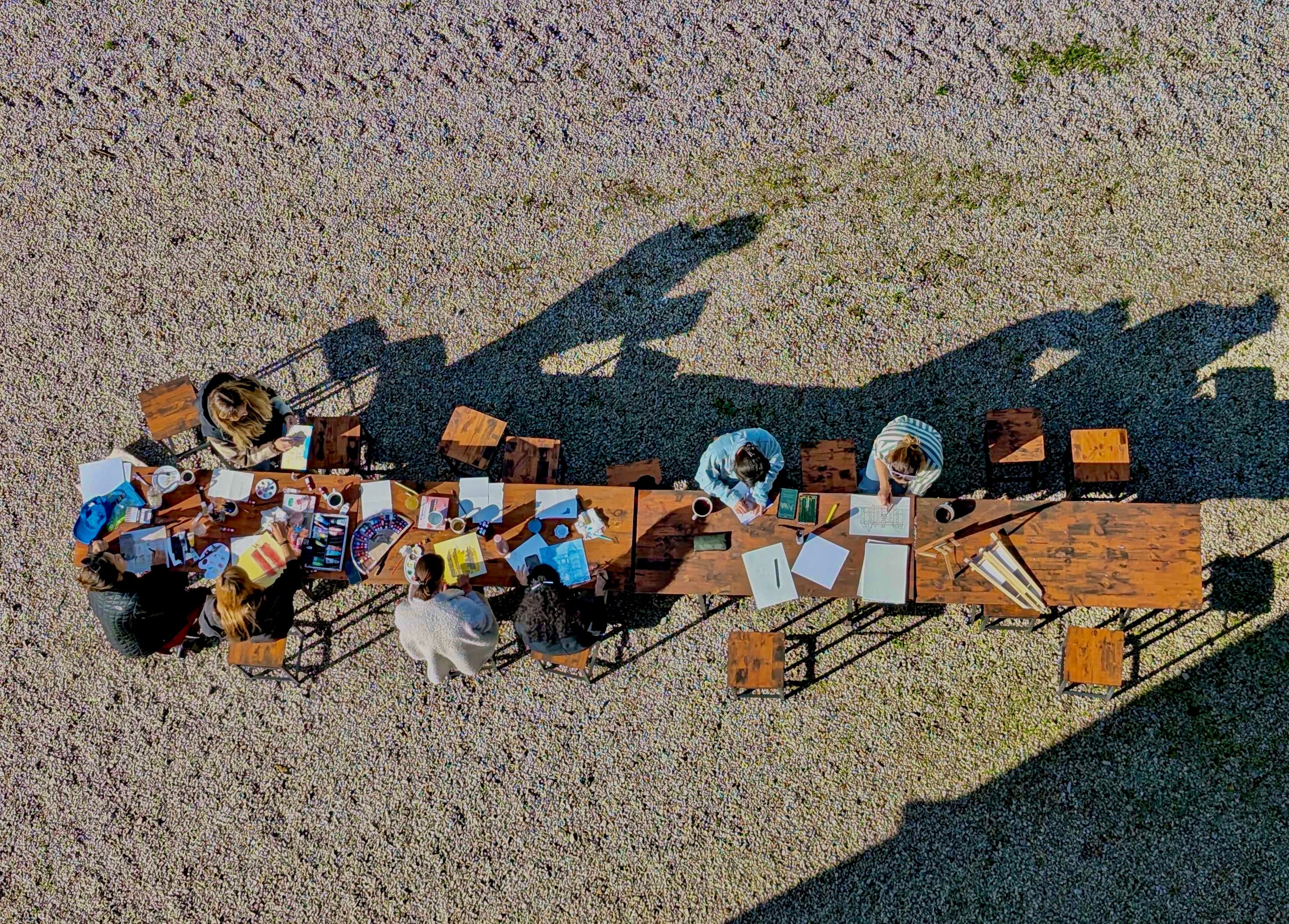 Aerial top-down view of a group working at a long wooden table on a gravel courtyard, notebooks, art supplies and coffee scattered across the table, with long sunlit shadows.