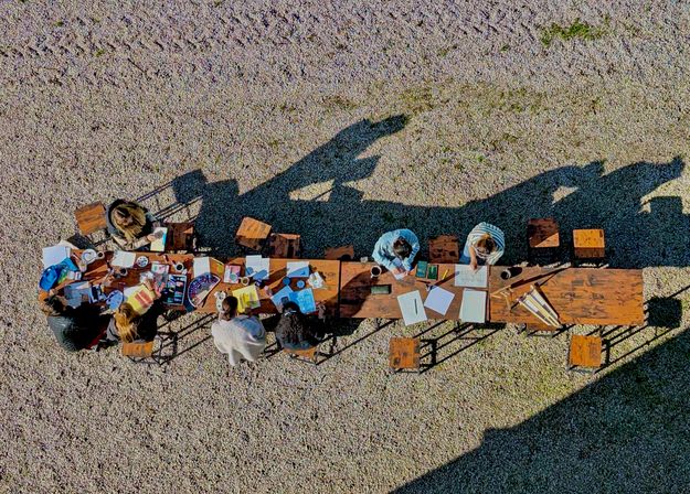 Aerial top-down view of a group working at a long wooden table on a gravel courtyard, notebooks, art supplies and coffee scattered across the table, with long sunlit shadows.
