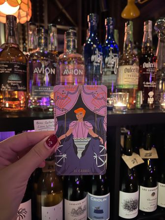 Hand with glossy red nails holding a colorful 'El Carro' tarot card in front of a candlelit bar shelf stocked with tequila and wine bottles.
