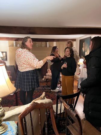 Cozy living room with a small group of adults in animated conversation, drinks in hand, under exposed wooden beams around a fireplace amid antique chairs, patterned rug and warm lamp lighting.