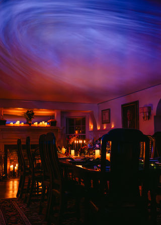 Cozy candlelit dining room at night — long wooden table set for dinner with flowers and candles, warm fireplace glow, and swirling blue ceiling light projection.