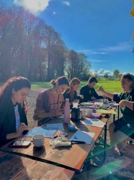 Group art session: five people painting and drawing at a long wooden picnic table in a sunny park meadow with tall trees and blue sky.
