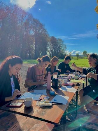 Group art session: five people painting and drawing at a long wooden picnic table in a sunny park meadow with tall trees and blue sky.