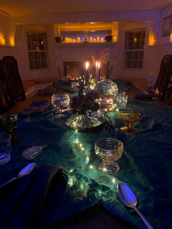 Candlelit dining table in a cozy living room: teal lace runner with twinkling fairy lights, crystal globe centerpiece, vintage goblets and spoons, and a warm fireplace glow.