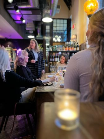 Group of women enjoying a wine tasting at a cozy urban wine bar, sitting around a wooden communal table with wine glasses, bottle-lined shelves, warm pendant lights and a candlelit ambiance.