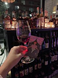 Hand holding a glass of red wine and an 'El Carro' tarot card in front of a candlelit shelf of wine and spirits in a cozy urban wine bar at night