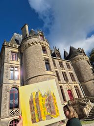 Plein-air scene: a person holds a sunlit bright-yellow watercolor of a historic stone chateau with round turrets, red-trim windows and ornate stairway under a blue sky with clouds.