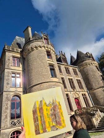 Plein-air scene: a person holds a sunlit bright-yellow watercolor of a historic stone chateau with round turrets, red-trim windows and ornate stairway under a blue sky with clouds.