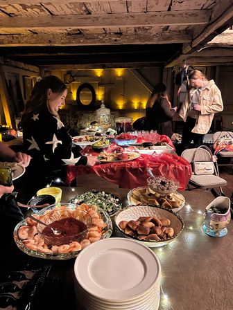 Rustic barn-style indoor buffet with shrimp cocktail, bread rolls, chips, salads and desserts on a red-draped table, guests serving themselves under warm string lights and exposed wooden beams.