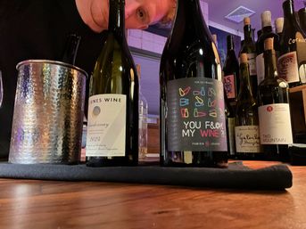 Close-up of wine bottles and a hammered metal spittoon on a wooden bar counter in a cozy wine-tasting room; playful black bottle label reads 'YOU F&%K MY WINE?!' with a person peeking behind the bottles.
