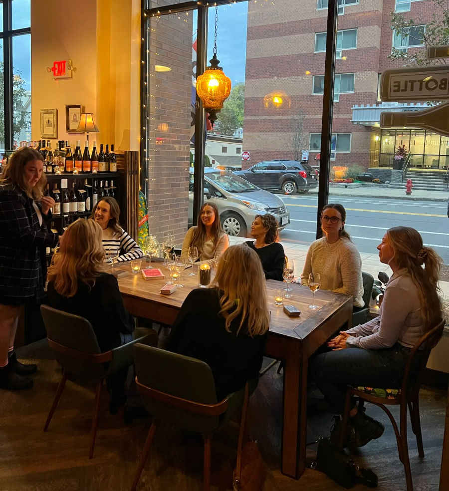 Group of friends enjoying a candlelit wine tasting at a cozy downtown wine bar, seated around a wooden table by large street-facing windows with warm pendant lighting.