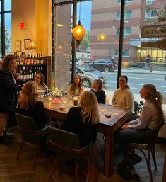 Group of friends enjoying a candlelit wine tasting at a cozy downtown wine bar, seated around a wooden table by large street-facing windows with warm pendant lighting.