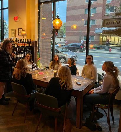 Group of friends enjoying a candlelit wine tasting at a cozy downtown wine bar, seated around a wooden table by large street-facing windows with warm pendant lighting.