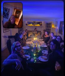 Festive candlelit dinner party in a cozy home dining room: a long table set with plates, candles, string lights and a floral centerpiece under blue ambient lighting, about a dozen guests seated around the table and a small selfie video-inset in the top corner.