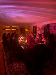 Candlelit dinner party with a group seated around a long table in a cozy home dining room, pink swirled ceiling light and mantel candles casting a warm ambient glow