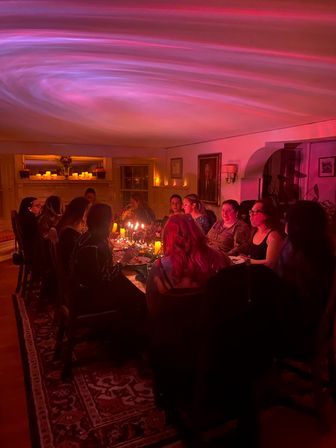 Candlelit dinner party with a group seated around a long table in a cozy home dining room, pink swirled ceiling light and mantel candles casting a warm ambient glow