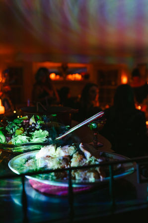 Colorful indoor dinner party buffet with a salad bowl and roasted chicken on plates, cocktail glass nearby, warm candlelit silhouettes and vibrant ceiling lights.