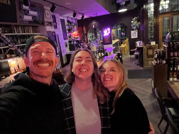 Three friends taking a selfie in a cozy, dimly lit wine bar with a neon sign, wooden bar, stools, and shelves of bottles.