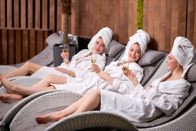 Three women in white robes and towel turbans relaxing on wicker lounge chairs in an indoor spa lounge, smiling and toasting champagne glasses.