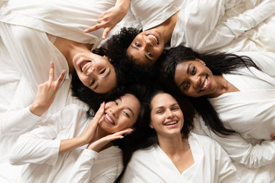 Five friends in white robes lying head-to-head on a bed, smiling and posing for a playful spa-day pajama party photo.