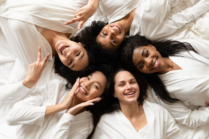 Five friends in white robes lying head-to-head on a bed, smiling and posing for a playful spa-day pajama party photo.