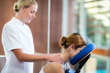 Smiling massage therapist giving a seated chair neck and shoulder massage to a woman resting on a blue face cradle in a bright modern spa