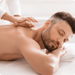 Relaxed man lying face-down with eyes closed receiving a shoulder massage at a bright wellness spa, therapist’s hands easing tension.