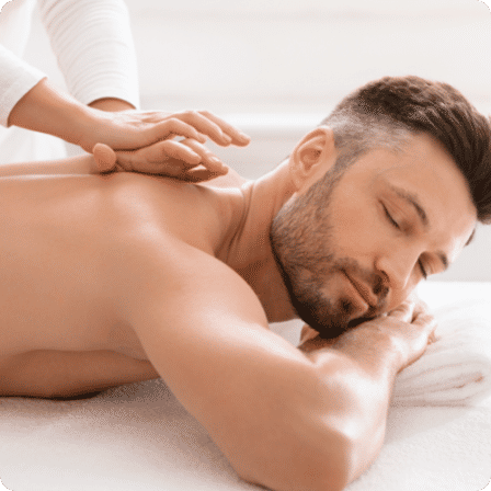 Relaxed man lying face-down with eyes closed receiving a shoulder massage at a bright wellness spa, therapist’s hands easing tension.
