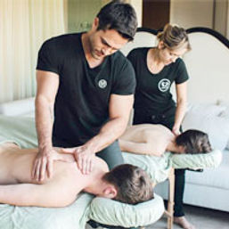 Two massage therapists in black shirts giving side-by-side back massages to clients on portable tables in a bright spa treatment room for relaxation and wellness.