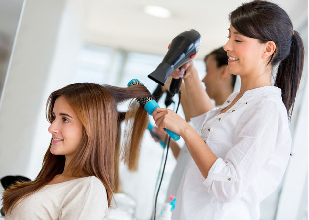 Smiling stylist blow-drying a client's long brown hair with a round brush and hair dryer in a bright salon