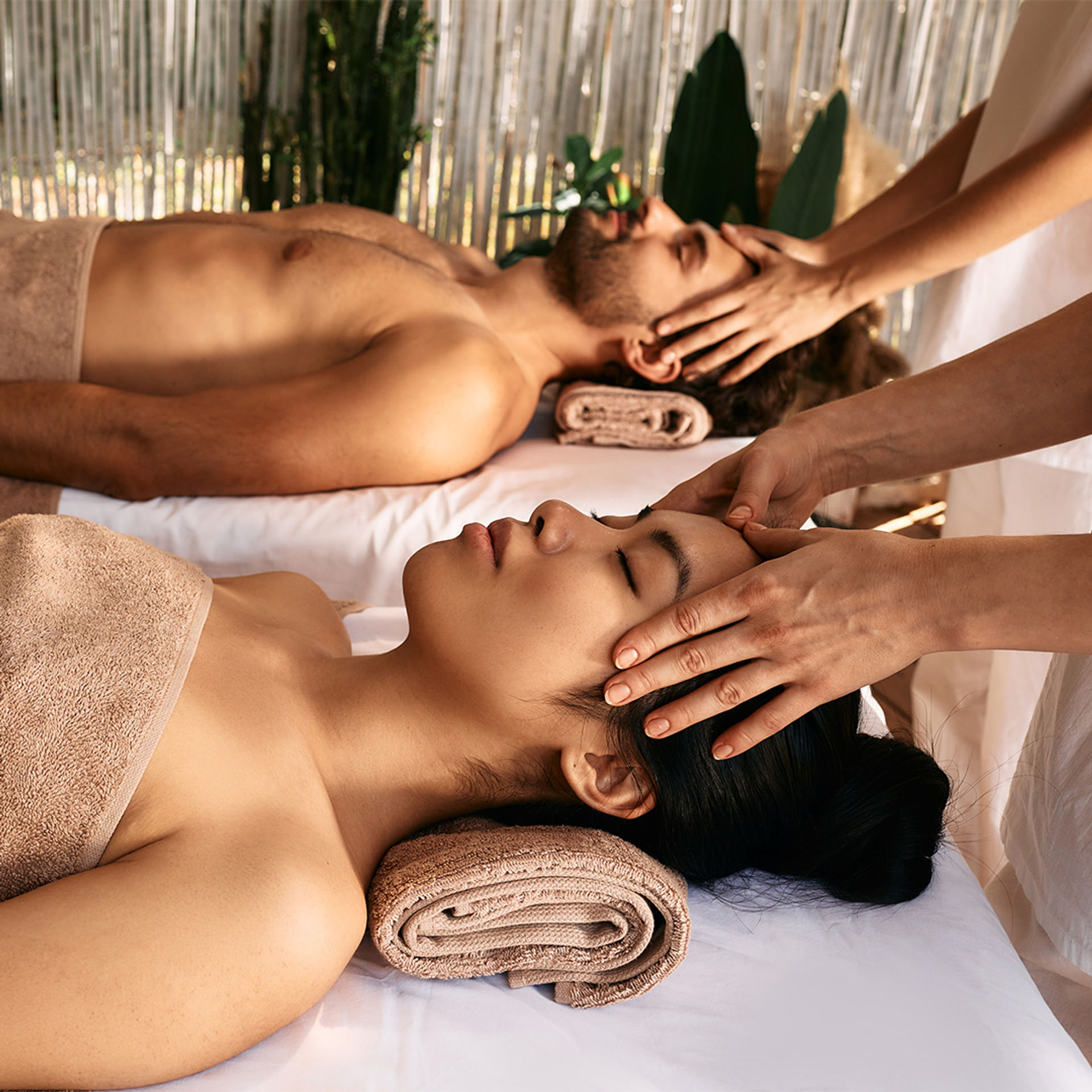 Couple receiving relaxing head and facial massages side-by-side on spa tables with rolled towels and tropical plants in a serene wellness spa setting.
